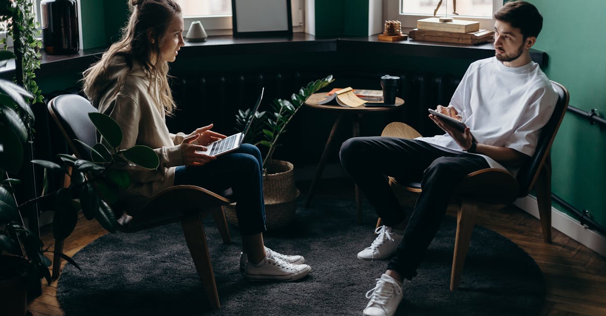 A man and woman engaged in conversation at a stylish, plant-filled home office with modern furniture and digital devices.